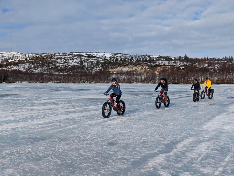 Gruppe von Fahrern, die mit Elektro-Fatbikes über einen zugefrorenen See bei Kirkenes fahren, mit schneebedeckten Hügeln im Hintergrund