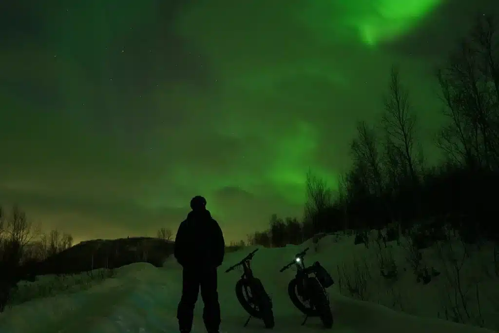 Person standing next to e-fatbikes under the Northern Lights during a winter night tour in Kirkenes, Northern Norway.