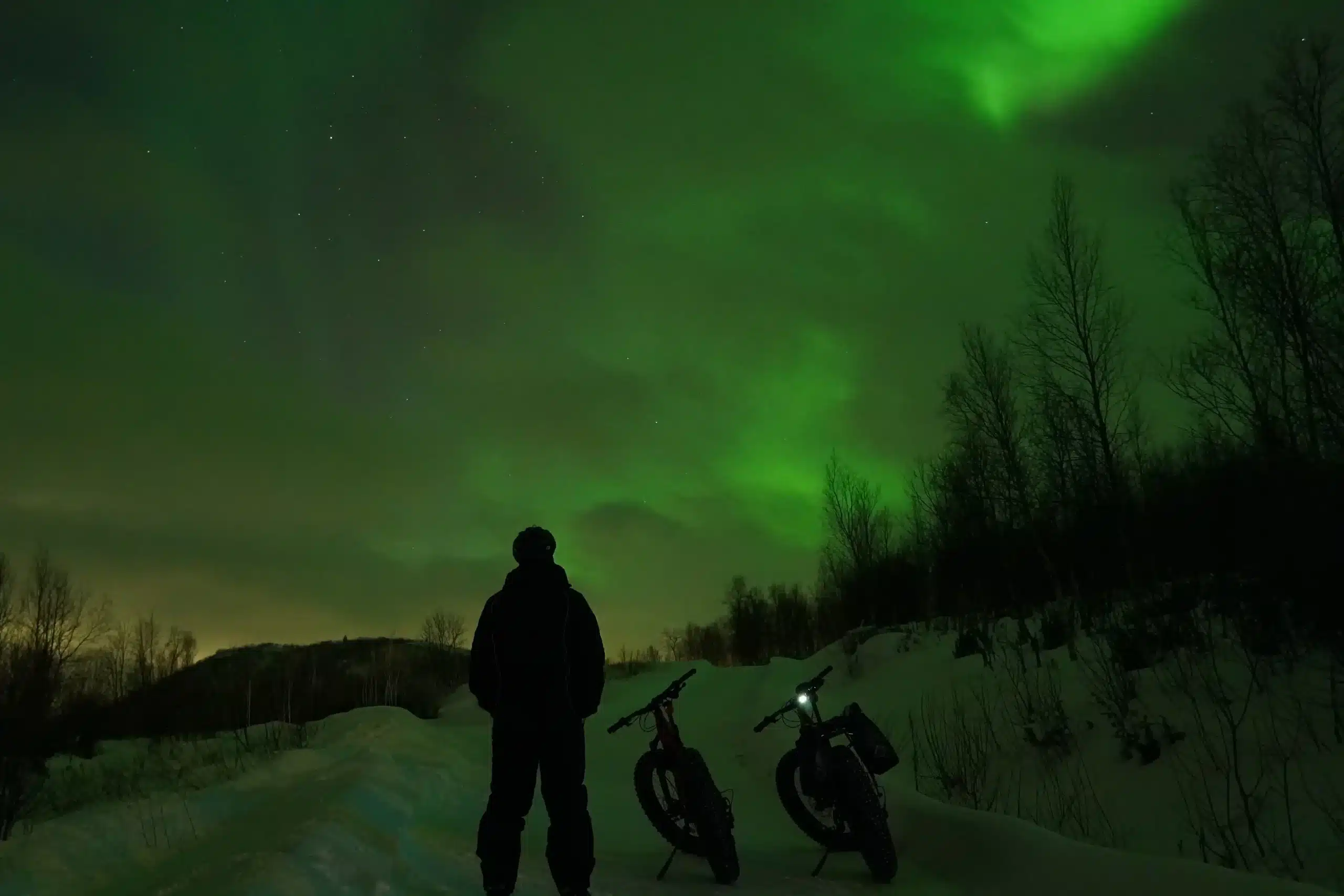 Person standing next to e-fatbikes under the Northern Lights during a winter night tour in Kirkenes, Northern Norway.