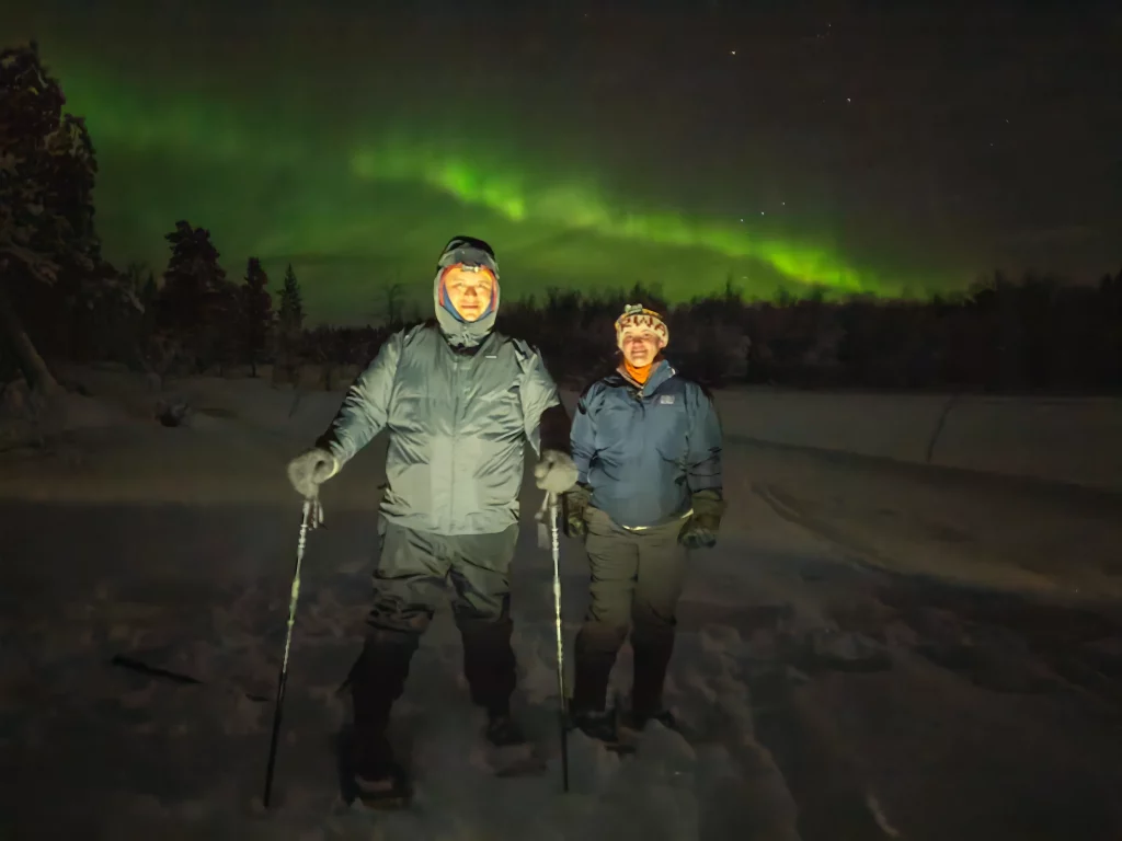 Two guests snowshoeing through deep snow near Kirkenes at night with green Northern Lights glowing above the winter forest.