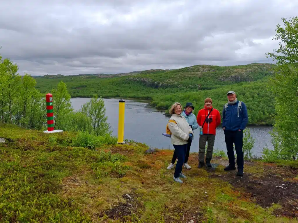 A small group of hikers standing beside the Norwegian and Russian border markers overlooking the Pasvik River near Kirkenes, with panoramic views across the Arctic border landscape.