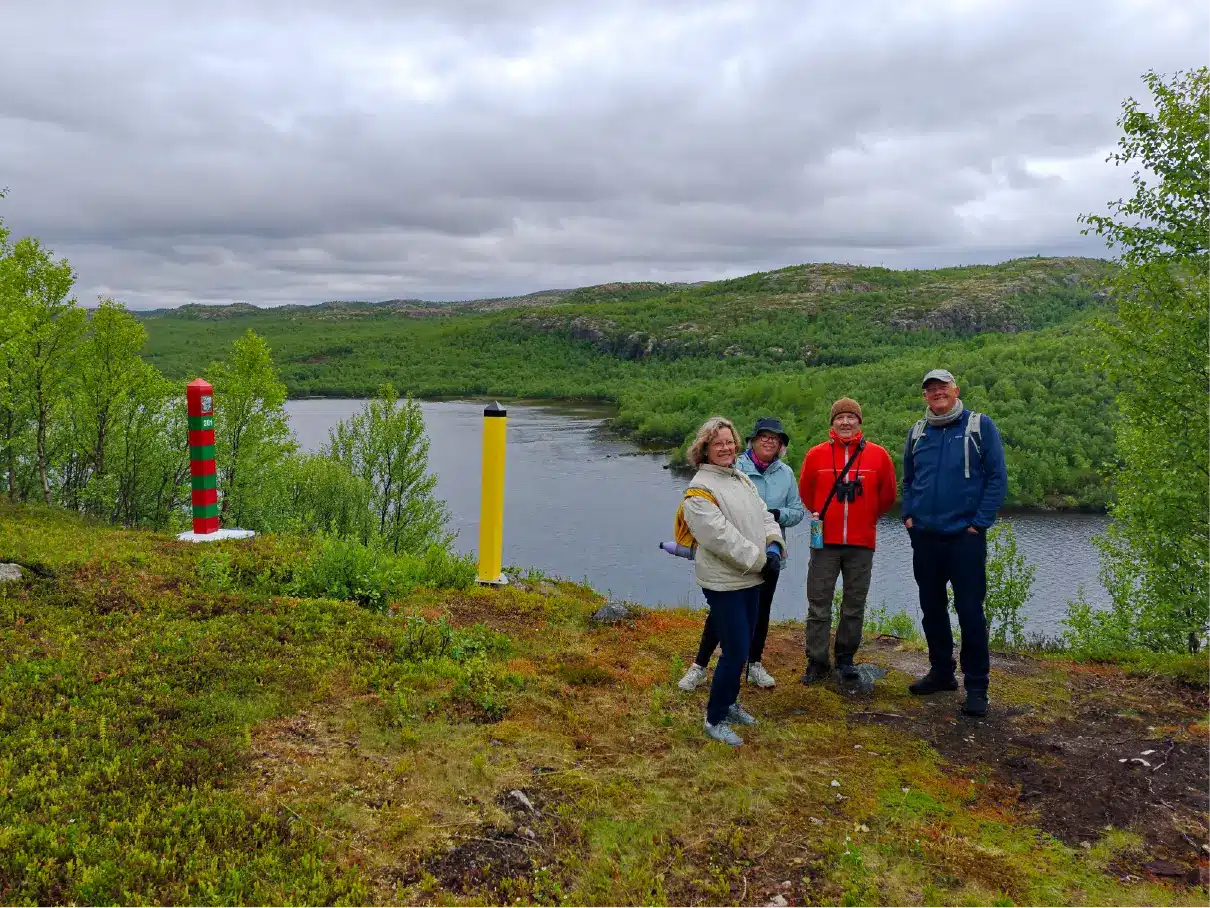 Eine kleine Gruppe von Wanderern steht neben den norwegischen und russischen Grenzsteinen und blickt auf den Grenzwasser Fluss Pasvik in der Nähe von Kirkenes, mit Panoramablick über die arktische Grenzlandschaft.