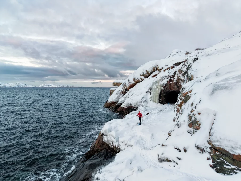 Luftaufnahme einer Person, die mit Schneeschuhen entlang verschneiter Küstenklippen in der Nähe einer Seehöhle und eines gefrorenen Wasserfalls an der Barentssee bei Kirkenes im arktischen Norwegen wandert.