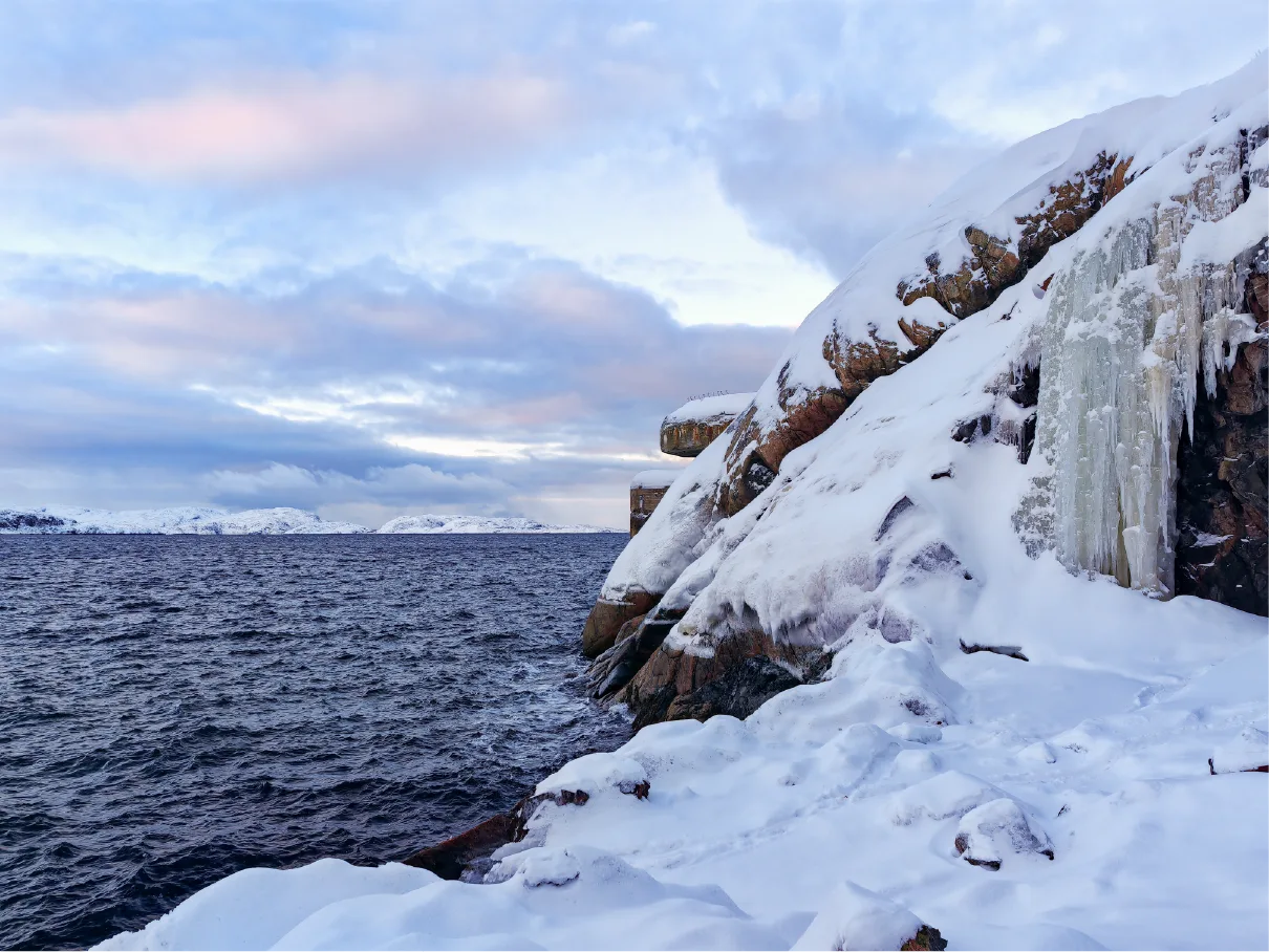 Schneebedeckte Küstenklippen mit einem gefrorenen Wasserfall und einem Torpedobatteriebunker aus dem Zweiten Weltkrieg mit Blick auf die Barentssee bei Kirkenes im arktischen Norwegen.