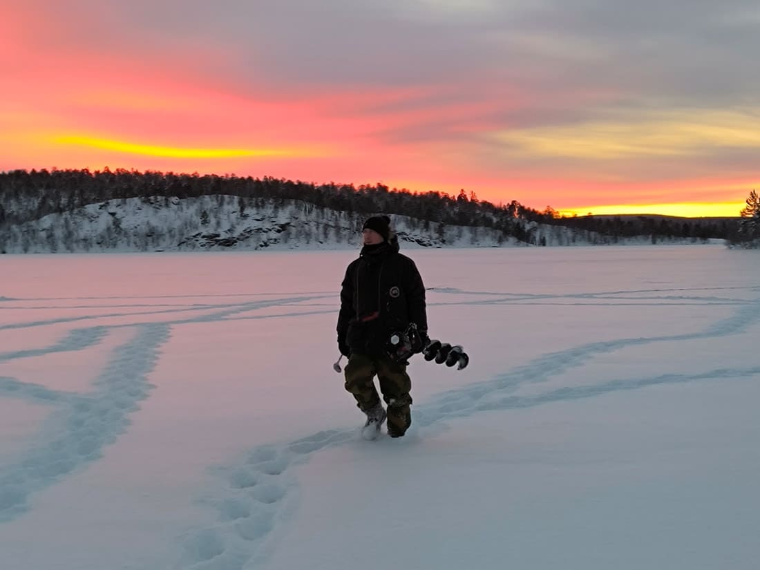 Man walking with ice drill across frozen lake to prepare for ice fishing trip near Kirkenes in Northern Norway