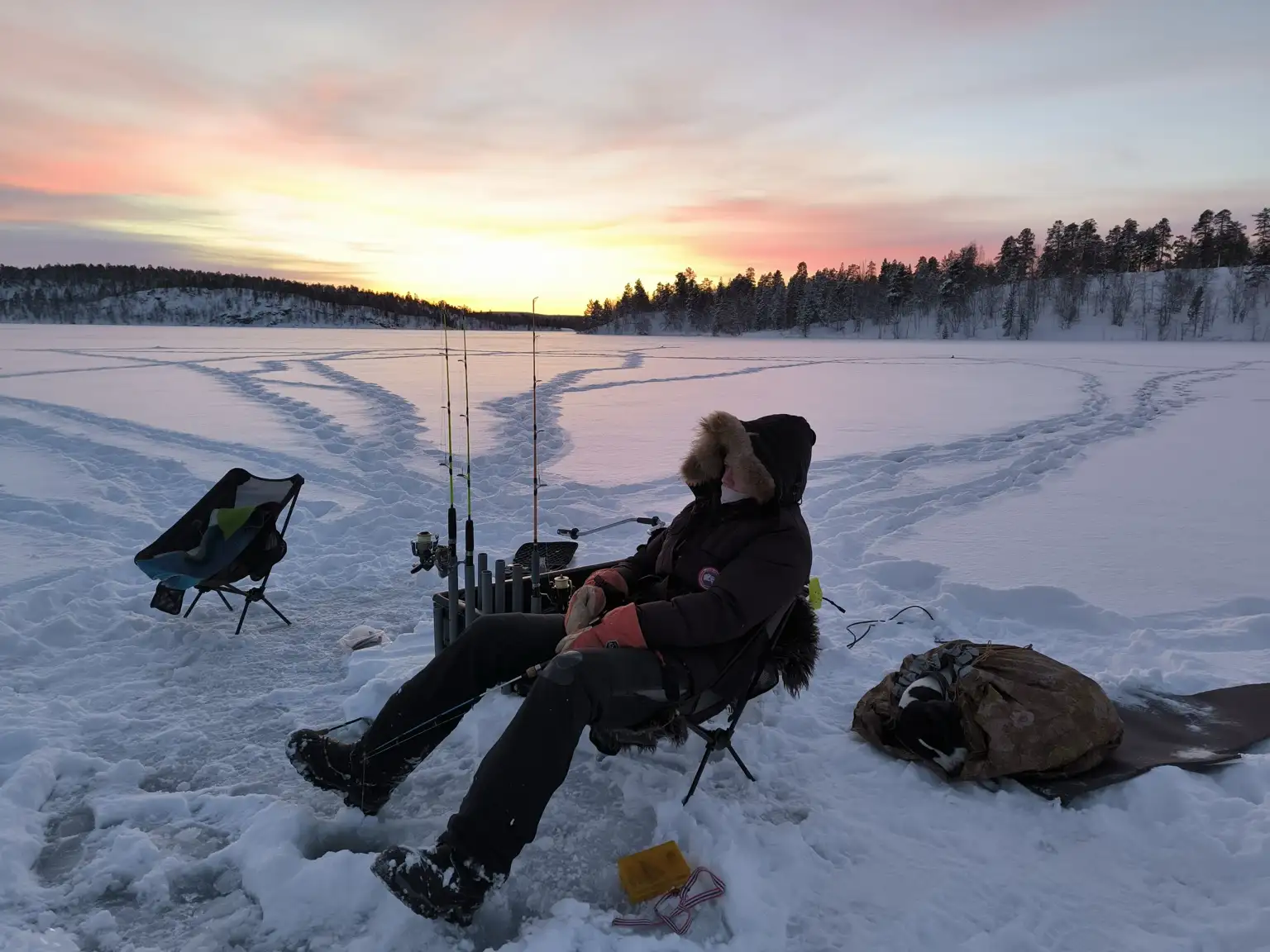 Person relaxing while ice fishing on a frozen lake during a colorful Arctic sunrise in Kirkenes, Norway.