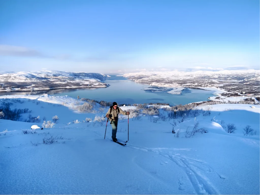 Skier touring across a snowy mountain plateau near Kirkenes with panoramic views over Jarfjord and snow covered hills under clear winter skies.