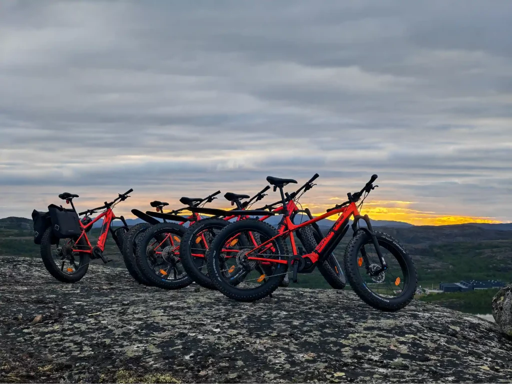 Five red e-fatbikes parked on a rocky summit overlooking the fjords and Arctic landscape near Kirkenes during the midnight sun.