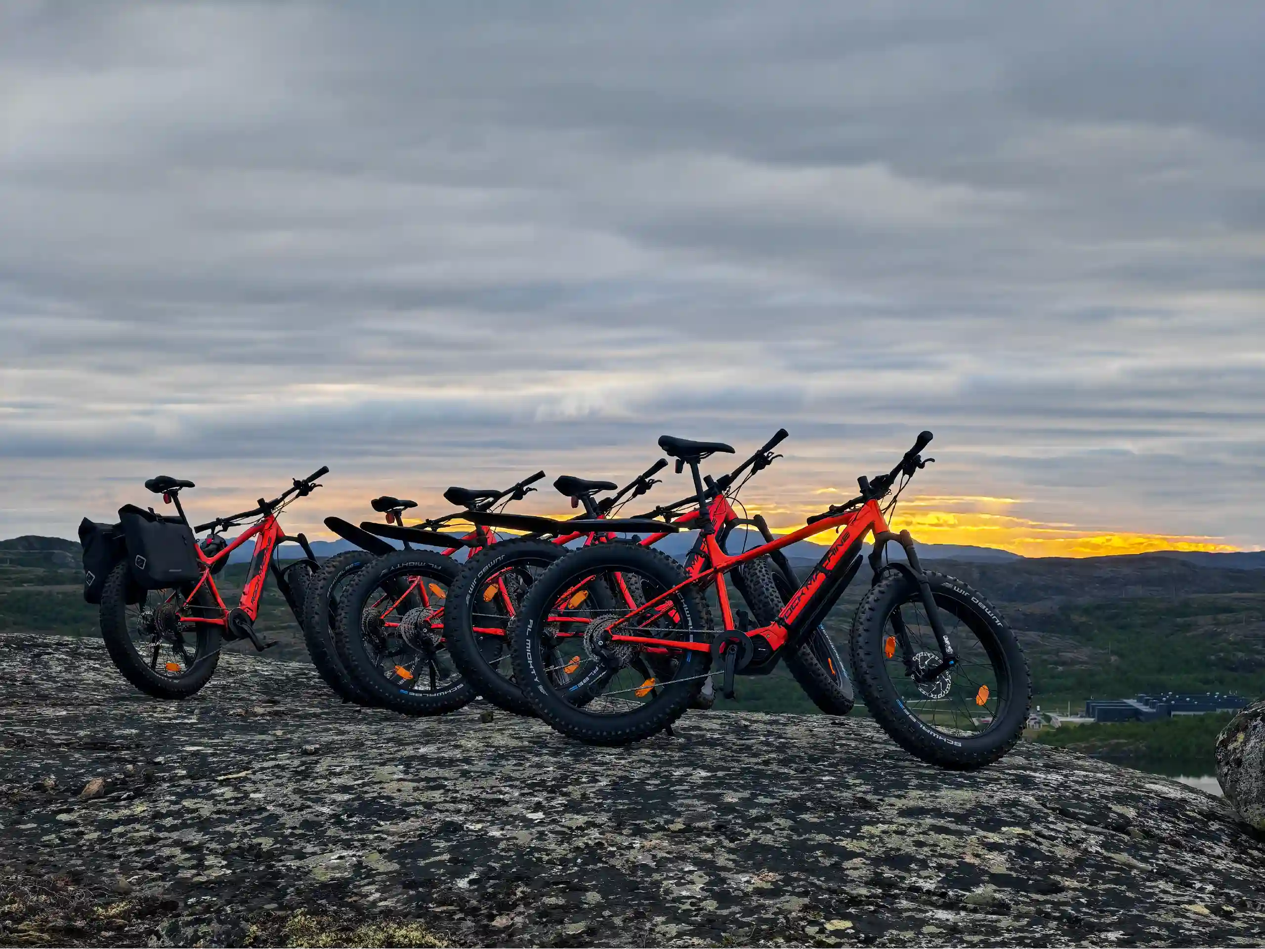 Five red e-fatbikes parked on a rocky summit overlooking the fjords and Arctic landscape near Kirkenes during the midnight sun.