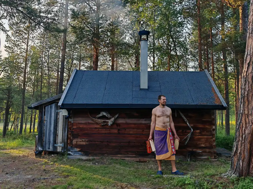 Man wrapped in a towel standing outside a traditional wood-fired sauna cabin in a pine forest at Namdalen Wilderness Lodge near Kirkenes, Northern Norway