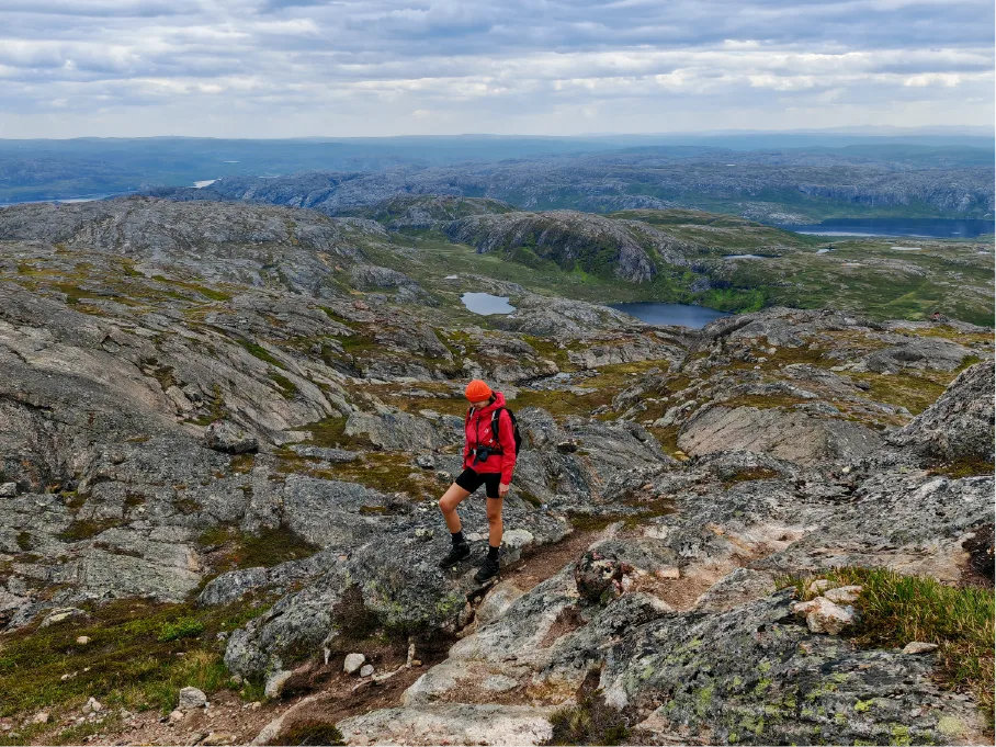 Hiker in a red jacket and orange cap standing on a rocky Arctic mountain ridge at Øretoppen near Kirkenes, overlooking lakes and vast tundra under cloudy skies.