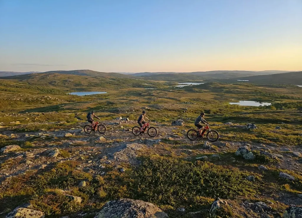 Three cyclists riding electric fat bikes across a rocky Arctic plateau near Kirkenes, surrounded by rolling hills and small lakes under the warm evening light.