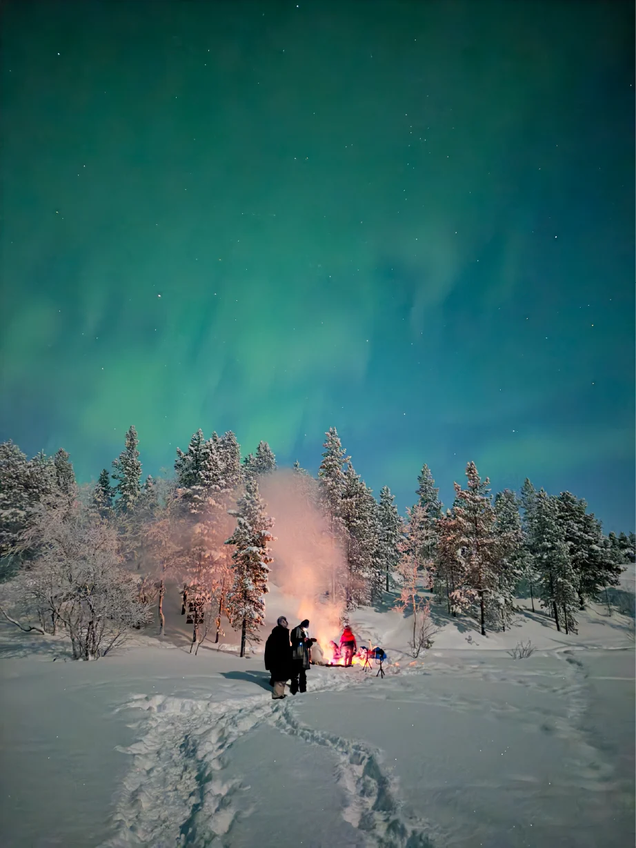 Leute haben sich um ein Lagerfeuer im verschneiten Wald unter den Nordlichtern bei Kirkenes versammelt.