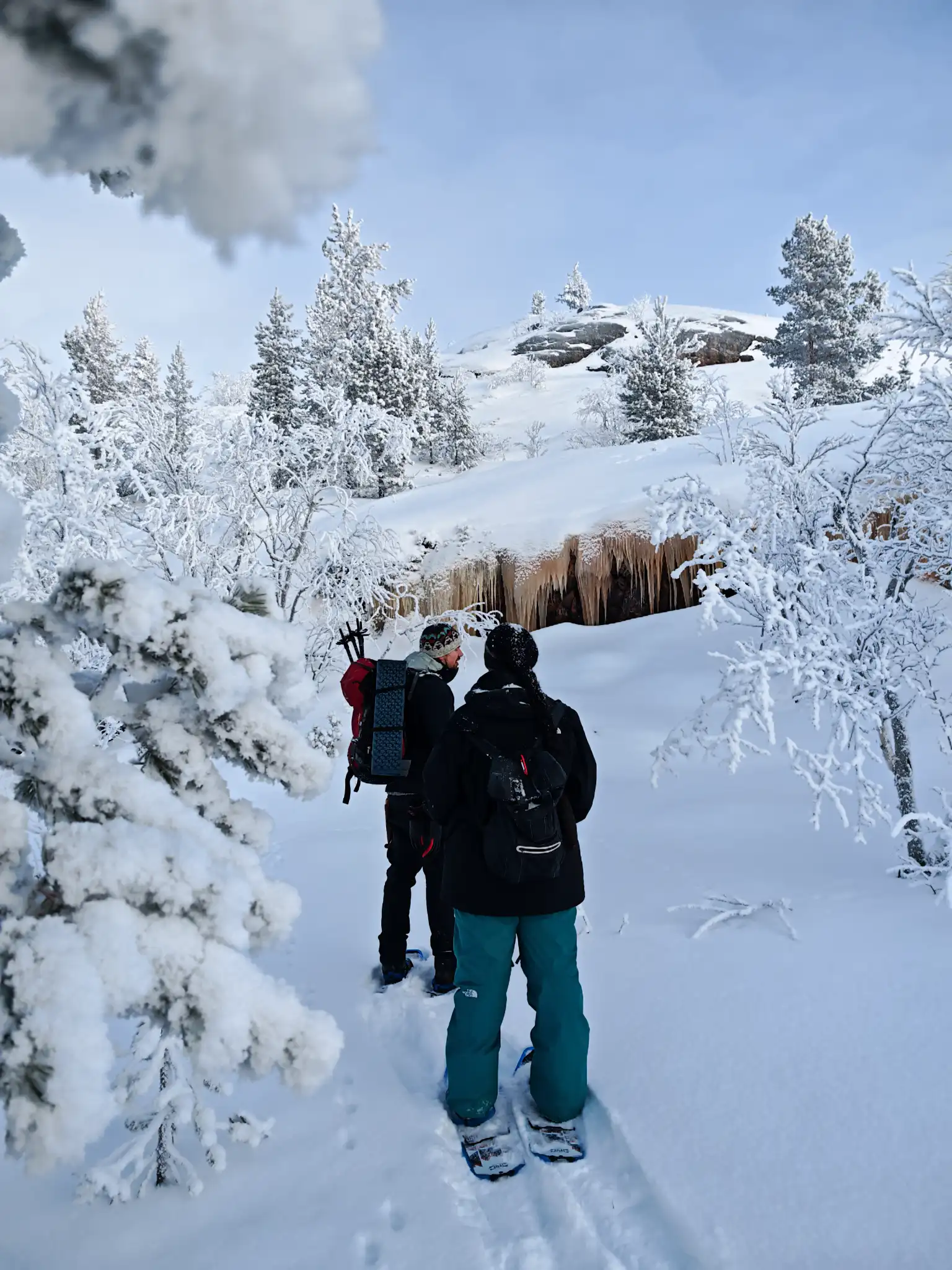 Zwei Leute wandern mit Schneeschuhen durch tiefen Schnee auf gefrorene Eiszapfen und eine felsige Anhöhe zu in einer Winterlandschaft entlang der norwegisch-russischen Grenze bei Kirkenes in Nordnorwegen