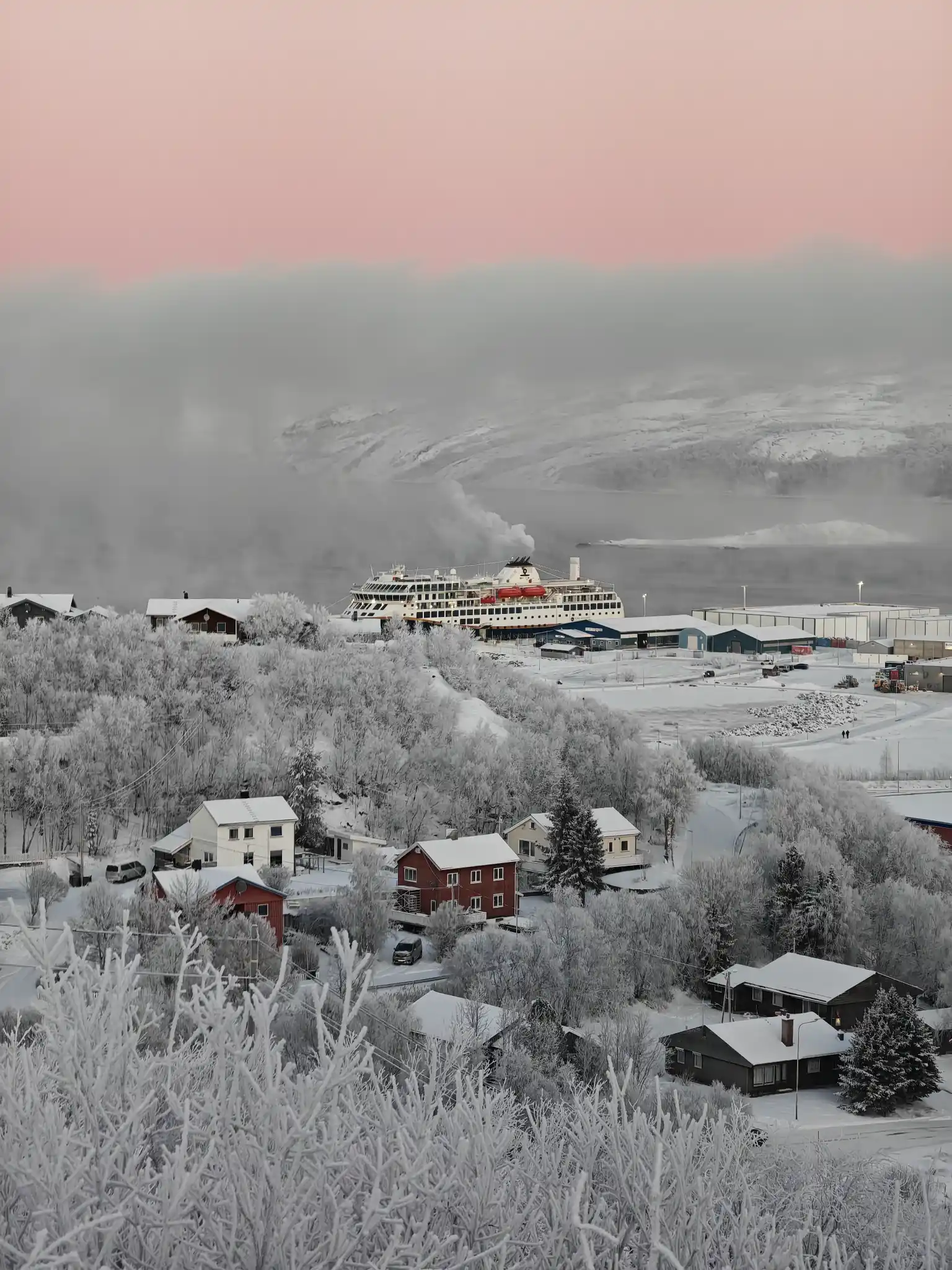 Schnee bedeckte Kirkenes mit einem Havila-Schiff im Hafen, umgeben von einer Winterlandschaft und einem zartrosa Himmel