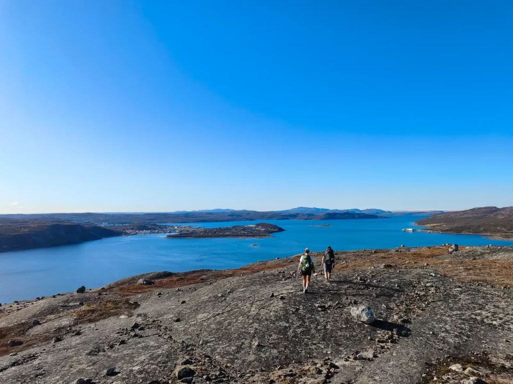 Two hikers walking along a bare rock ridge above Kirkenes, with the deep blue Varangerfjord and the town spread out in the distance under the midnight sun in May