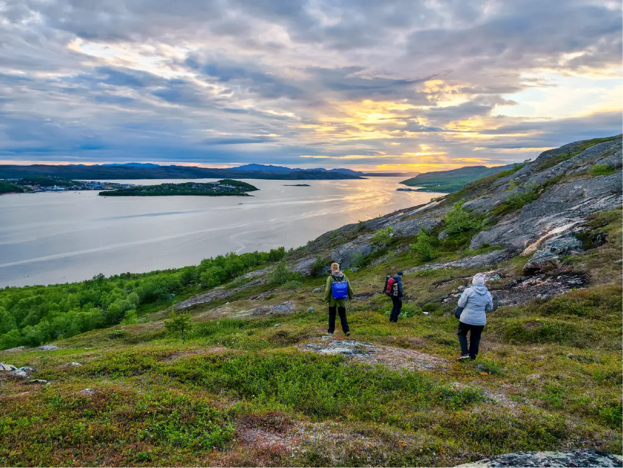 Guests hiking across the hills above Kirkenes during the midnight sun, overlooking Bøkfjorden, the town of Kirkenes, and the surrounding Arctic landscape under a colorful summer sky.