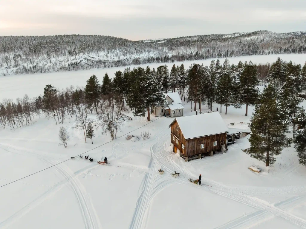 Hundeschlitten-Teams passieren die Namdalen Wilderness Lodge in einer schneereichen Winterlandschaft an einem zugefrorenen See im Pasvik-Tal in der Nähe von Kirkenes.