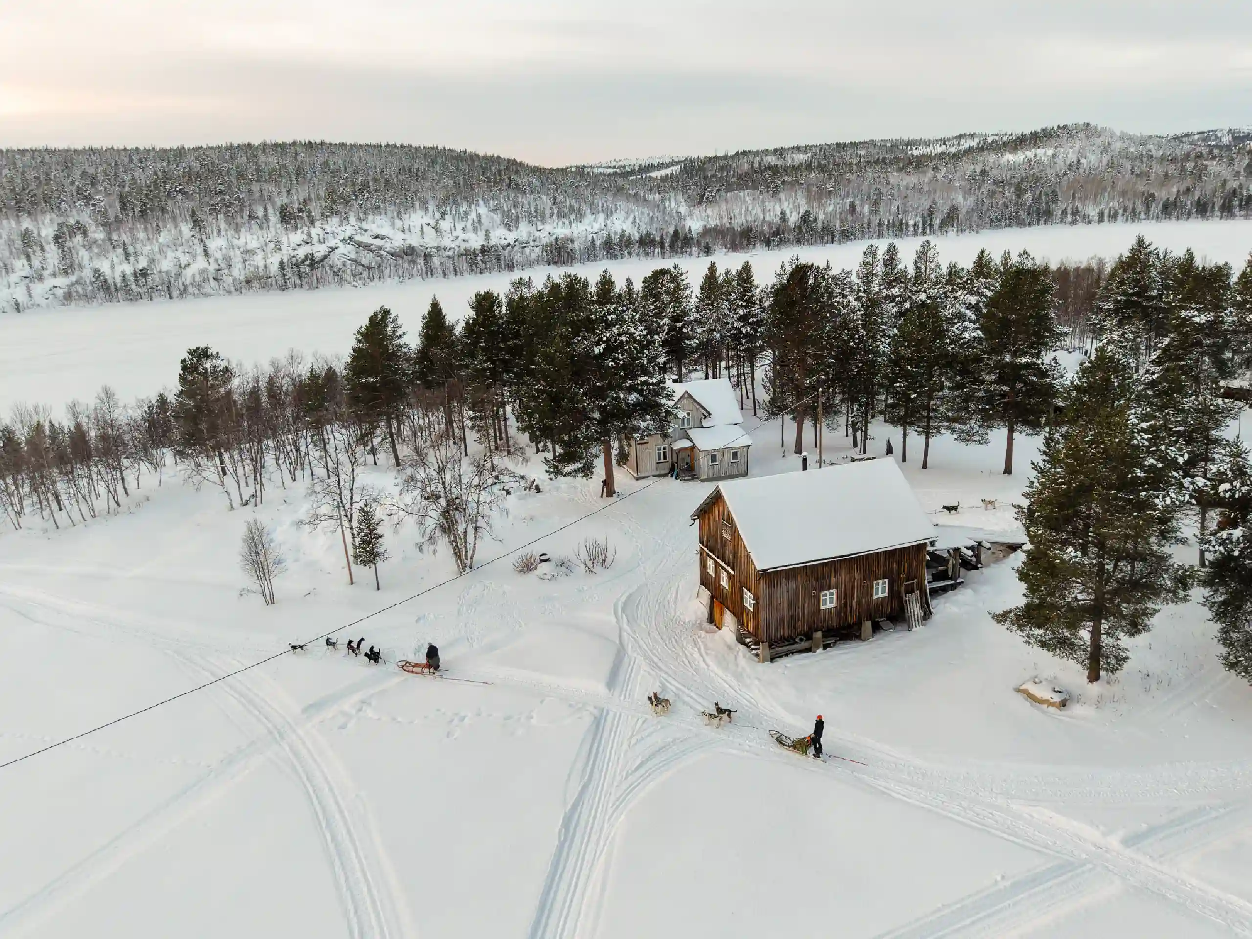Hundeschlitten-Teams passieren die Namdalen Wilderness Lodge in einer schneereichen Winterlandschaft an einem zugefrorenen See im Pasvik-Tal in der Nähe von Kirkenes.