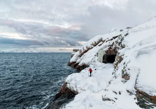 Aerial view of a person snowshoeing along snowy coastal cliffs near a sea cave and frozen waterfall on the Barents Sea near Kirkenes in Arctic Norway.