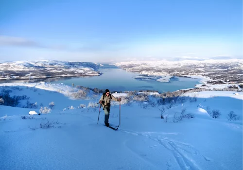 Skier touring across a snowy mountain plateau near Kirkenes with panoramic views over Jarfjord and snow covered hills under clear winter skies.