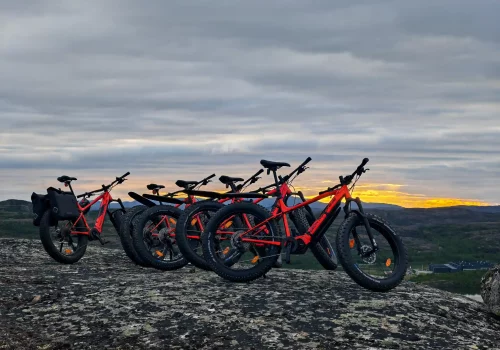 Five red e-fatbikes parked on a rocky summit overlooking the fjords and Arctic landscape near Kirkenes during the midnight sun.
