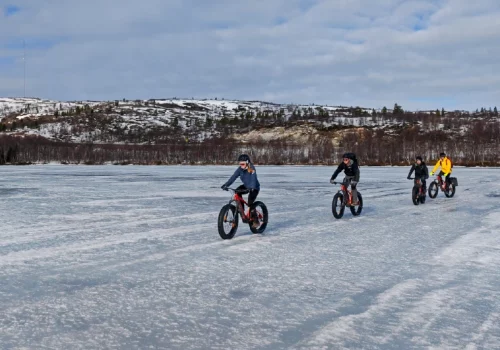 Group of riders cycling with electric fatbikes across a frozen lake near Kirkenes with snowy hills in the background
