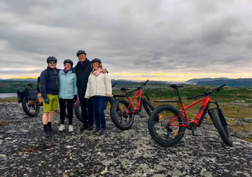 Four guests posing with red e-fatbikes at a scenic viewpoint overlooking the fjords and Arctic landscape near Kirkenes during a guided e-fatbike tour.
