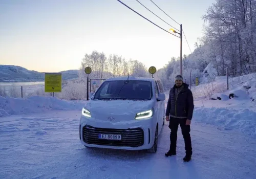 Kirkenes Tours guide standing beside a white Toyota ProAce Verso minivan on a snowy road near Kirkenes, Northern Norway, surrounded by frost covered trees.