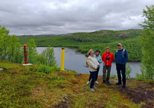 A small group of hikers standing beside the Norwegian and Russian border markers overlooking the Pasvik River near Kirkenes, with panoramic views across the Arctic border landscape.