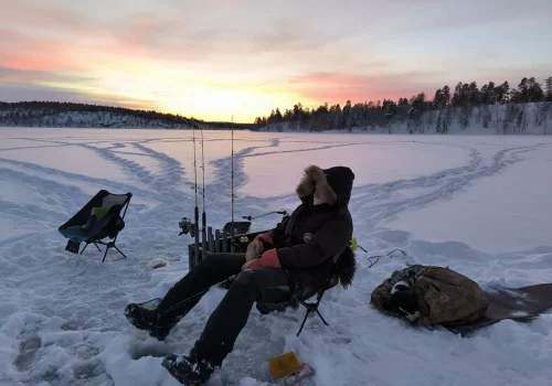 Person relaxing while ice fishing on a frozen lake during a colorful Arctic sunrise in Kirkenes, Norway.
