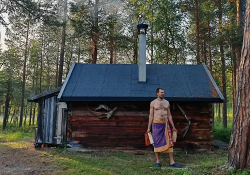 Man wrapped in a towel standing outside a traditional wood-fired sauna cabin in a pine forest at Namdalen Wilderness Lodge near Kirkenes, Northern Norway