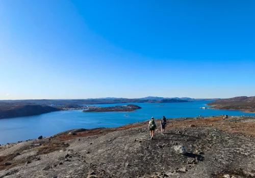 Two hikers walking along a bare rock ridge above Kirkenes, with the deep blue Varangerfjord and the town spread out in the distance under the midnight sun in May