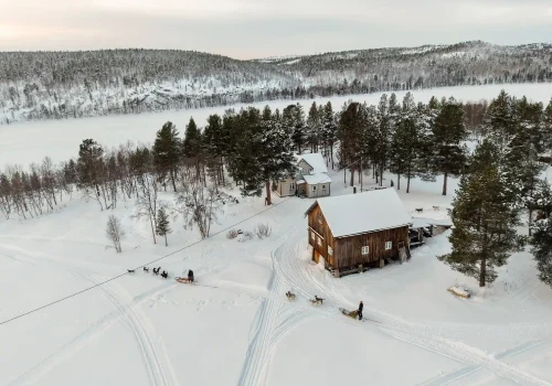Dog sled teams passing Namdalen Wilderness Lodge in a snowy winter landscape beside a frozen lake in the Pasvik Valley near Kirkenes.