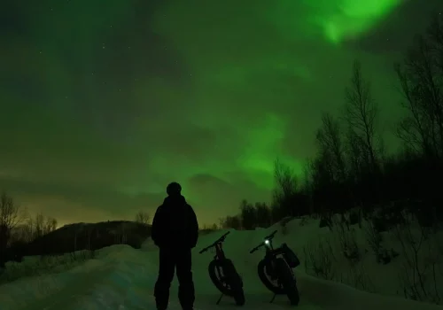 Person standing next to e-fatbikes under the Northern Lights during a winter night tour in Kirkenes, Northern Norway.