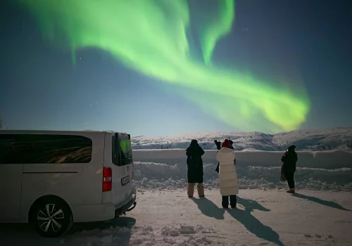 Gäste beobachten und fotografieren lebhafte grüne Nordlichter über schneebedeckten arktischen Bergen in der Nähe von Kirkenes, während nachts ein Tourbus am Straßenrand parkt.
