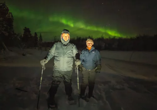 Two guests snowshoeing through deep snow near Kirkenes at night with green Northern Lights glowing above the winter forest.