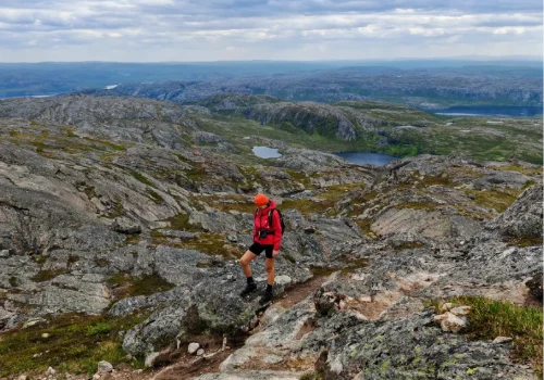 Hiker in a red jacket and orange cap standing on a rocky Arctic mountain ridge at Øretoppen near Kirkenes, overlooking lakes and vast tundra under cloudy skies.