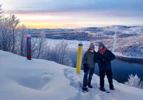 Two guests wearing snowshoes stand beside the Norwegian and Russian border markers overlooking the Pasvik River near Kirkenes, with snow covered hills and a colorful winter sky in the background.